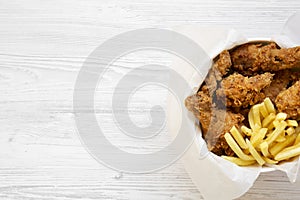 Fast food: fried chicken drumsticks, spicy wings, French fries and tender strips in paper box over white wooden surface, view from
