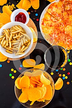 Fast food and unhealthy eating concept - close up of pizza,, french fries and potato chips and candies on wooden table top view