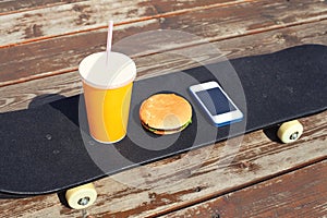Fast food and unhealthy eating concept - juice cup, hamburger, smartphone on skateboard