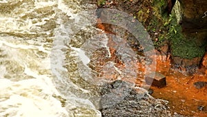 Fast full-flowing foamy water between sandstone rocks, orange sediments on dirty bank. Deep riverbed hewed into rock