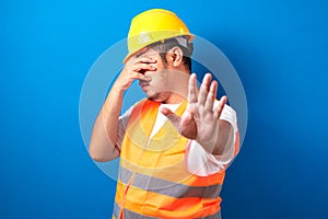 Fat asian construction worker man wearing uniform and helmet over isolated blue background covering eyes with hand