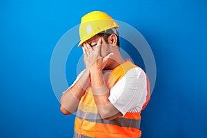 Fat asian construction worker man wearing uniform and helmet over isolated blue background covering eyes with hand