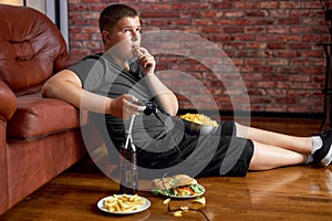 Fat boy sitting on floor in living room, side view. Overweight caucasian child at home