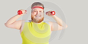 The fat funny man with a beard with dumbbells on a gray background.