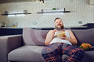 Fat funny man in pajamas eating a burger at home.