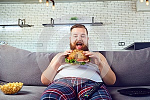 Fat funny man in pajamas eating a burger on the sofa at home.