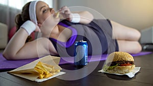 Fat lazy girl in sportswear eating junk food instead of training on yoga mat