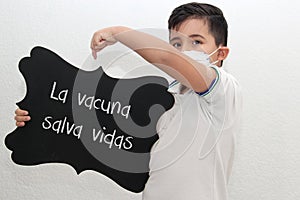 Fat little Hispanic boy with face mask and school uniform shirt shows his recently vaccinated arm against Covid-19 with a black si