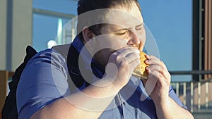 Fat male student eating high-calorie burger outdoors, fast food and overweight