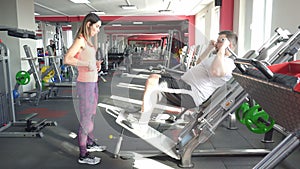 A fat man with a beard performs exercises on a calf leg simulator under the supervision of a girl trainer in a modern