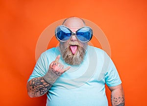 Fat man with beard, tattoos and sunglasses makes the gesture of the horns