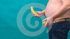 Fat man with a big belly holds a green apple and a banana in his hands. The concept of healthy eating and losing weight