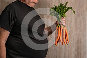 A fat man with a big belly humorously holds a bunch of carrots in his hand as a sign of a trend towards weight loss