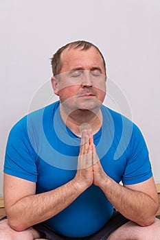 A fat man in a blue t-shirt is doing yoga, sitting on a fitness Mat and relaxing with his eyes closed and his hands