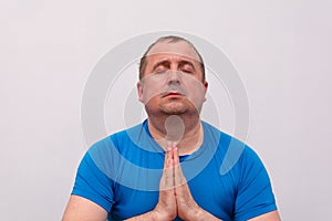 A fat man in a blue t-shirt is doing yoga, sitting on a fitness Mat and relaxing with his eyes closed and his hands clasped