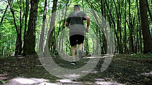 A fat man in a cap and shorts runs along a forest path in the forest in the summer. The concept of running for losing