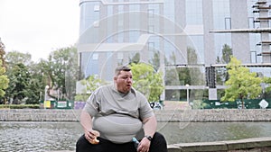 A fat man eats a hamburger and drinks water while exercising in the park. The problem of overweight in a young person