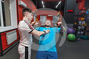 Fat man in a gym is diligently doing pull-ups under the guidance of a personal trainer. Overweight TRX