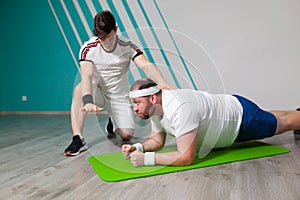 Fat man looks exhausted while doing exercises standing in bar on the mat in the fitness gym under strict control of