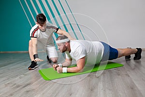 Fat man looks exhausted while doing exercises standing in bar on the mat in the fitness gym under strict control of