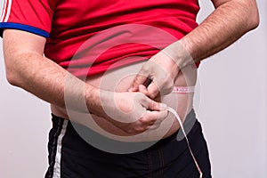 A fat man in a red t-shirt and black shorts measures his stomach with a centimeter tape, home fitness training