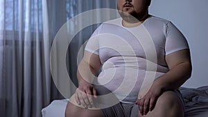 Fat man sitting on bed, weight gain due to sedentary lifestyle, hormonal disease