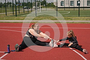 Fat obese mother and skinny daughter go in for sports together doing stretching exercises sitting on the sports surface of ground.