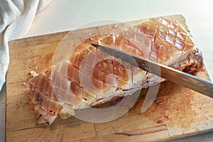 Fat rind of a piece of raw seasoned pork meat is incised with a kitchen knife on a cutting board, cooking preparation for a