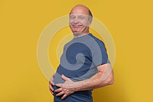 Fat senior man touching his big abdomen with hands after well eating.