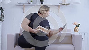 A fat woman lying on a couch reading a book and checking her phone at home