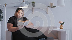 A fat woman lying on a couch and reading a book at home