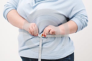 A fat woman measures her waist with a measuring tape in casual clothing on a white background.