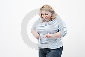 A fat woman measures her waist with a measuring tape in casual clothing on a white background.