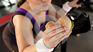 Fat woman riding stationary bike and eating fatty burger in gym, unhealthy diet