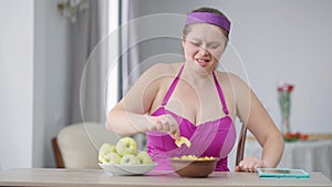 Fat young Caucasian woman breaking free from measuring tape on hands eating chips making strength gesture smiling