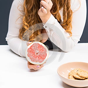 Fat young woman holding a fresh grapefruit in hand standing leaned on a kitchen table at home with long curly blond hair