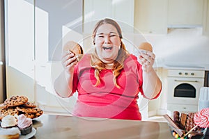 Fat young woman in kitchen sitting and eating junk food. Happy excited plus size model laughing. Holding burgers in