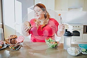 Fat young woman in kitchen sitting and eating junk food. Holding burger and tomato on fork. Regret and unhappiness. Body