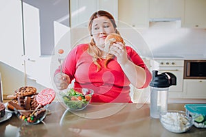 Fat young woman in kitchen sitting and eating junk food. Plus size model has unhealthy eating. Salad bowl on table. Body