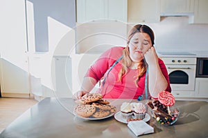 Fat young woman in kitchen sitting and eating sweet food. Bored plus size model look at pancakes and sweets on table