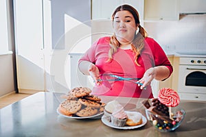 Fat young woman in kitchen sitting and eating sweet food. Soft tape measure around waist and in hands. Plus size model