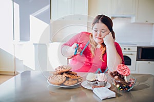 Fat young woman in kitchen sitting and eating sweet food. Upset plus size model look at cookies. Blue soft tape measure
