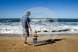 Father and 2 year old son facing ocean waves at the beach from b