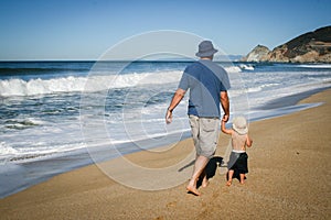 Father and 2 year old son walking along the sand at the beach in