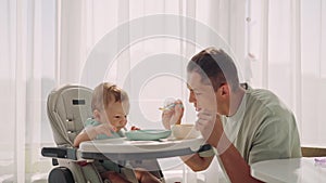 Father acting Mom feeding his son baby 1 on chair in the house. Father's day