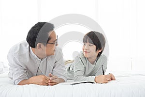 Father and Asian boy writing on book,lying on bed at home, Dad and son spending quality time together