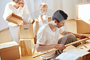 Father assembling furniture in living room of new apartment, mother with son and pile of moving boxes on background