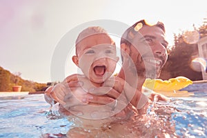 Father With Baby Daughter Having Fun On Summer Vacation Splashing In Outdoor Swimming Pool