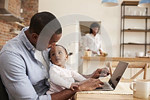 Father And Baby Daughter Use Laptop As Mother Prepares Meal