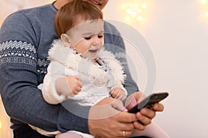 Father and baby use smartphone, they play and watch device screen, home interior decorated with holiday lights and gifts